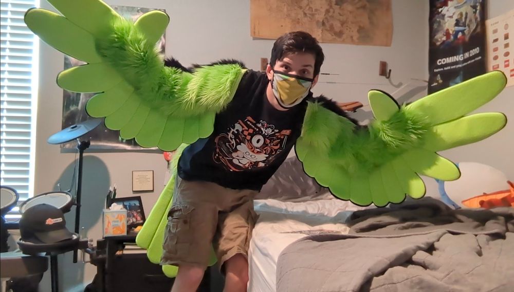 Real life photo of Sam wearing a pair of lime green wings over his arms. They are black on the back side. Also wearing a like green bird tail. Also have a yellow beak bird mask on his face.