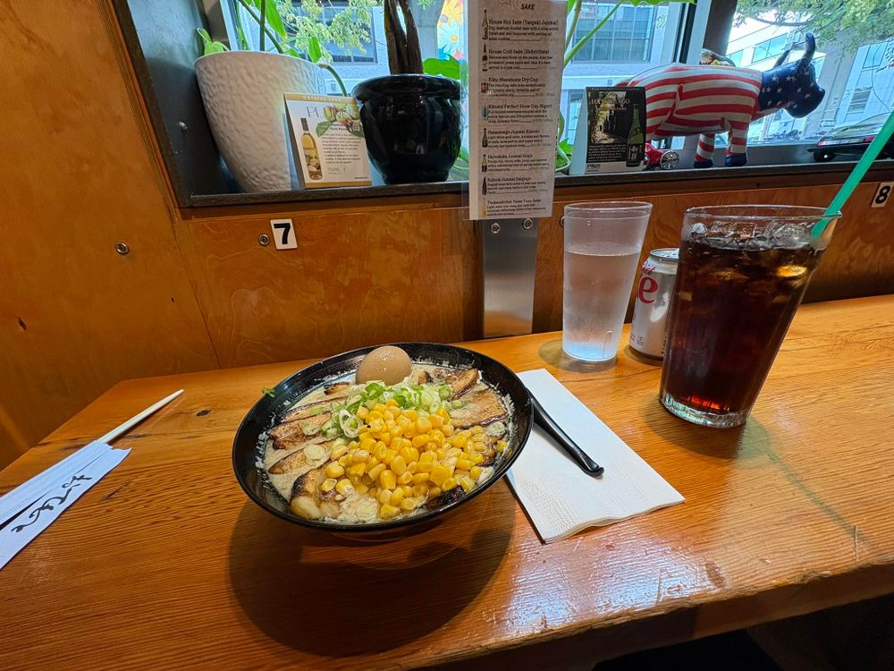 A seat at a cozy little ramen shop under a bridge. There's a bowl of tonkotsu ramen. You can see corn, scallions, a soft boiled egg, and plenty of pork belly on the surface. Next to it is a Diet Coke and a glass of ice water. It's appetizing.