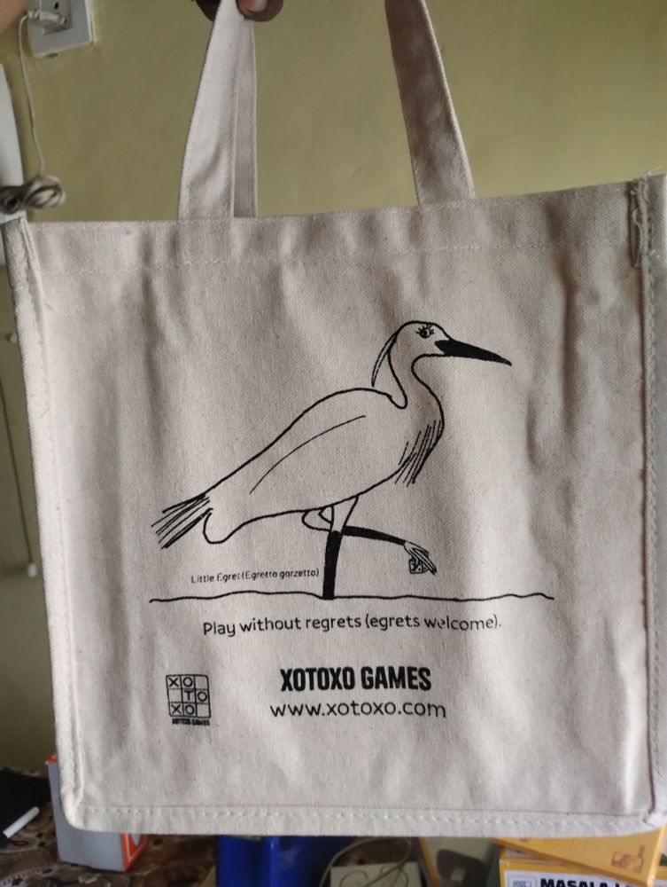 White canvas tote bag with a caricature of an egret holding a die on it. Caption reads "play without regrets (egrets welcome)". 
