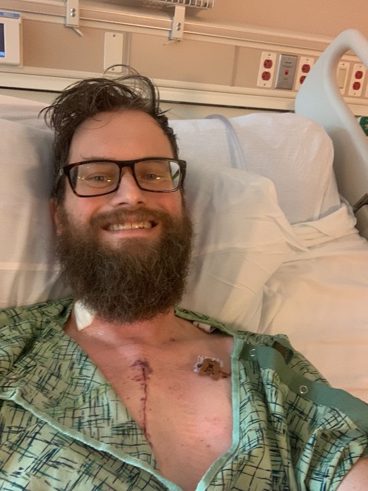 A man with a beard and glasses smiling on a hospital bed

He has a fresh scar down the middle of his chest and another on on his left shoulder 