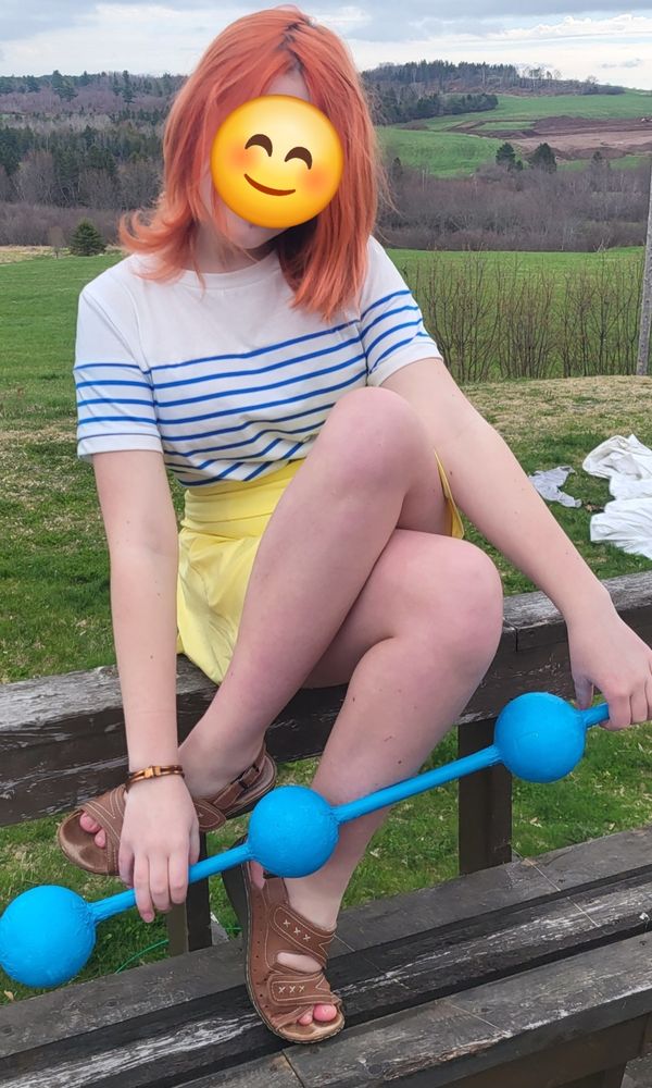 A young girl with orange hair dressed as Nami from One Piece sitting on her deck with Nami's weapon, the Climatact, a blue stick with three balls on it.