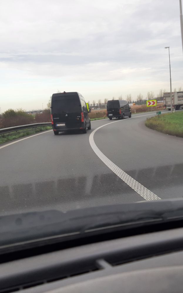 Zwei schwarze Transporter fahren auf einer kurvigen Autobahnauffahrt bei bedecktem Himmel, aufgenommen aus einem dahinter folgenden Fahrzeug heraus.

