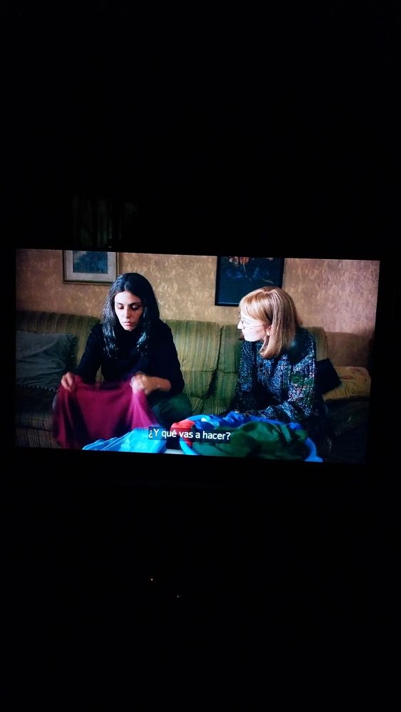 Dos mujeres, una morena y otra rubia, están sentadas en un sofá verde. La morena, Lila (Irene Maiolino), está doblando ropa y mira hacia abajo. La rubia, Lenù (Alice Rorhwacher) está mirando a Lila.