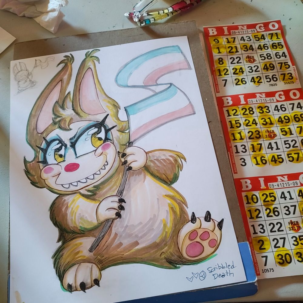 photo of a drawn next to a bingo card.

The drawing is the Labubu brown The Monster Pop Mart collectable doll, holding a trans flag. The monster has more colorful drag makeup, blue eyeliner ,bright red nose, thick thick pointy eyelashes and dotted rose circle cheeks.  Soft brown fluffy fur.

there's also a small thumbnail sketch at the top of the page