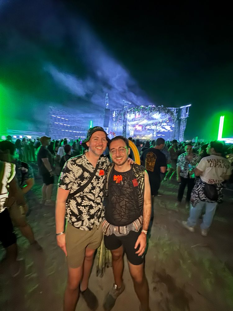 A nighttime music festival scene with two men posing near the front of a large crowd. Both are dressed in themed rave outfits; one wears a geometric tank and a fabric belt with patterns, while the other has a black floral shirt and khaki shorts. The air is filled with green and blue lights and smoke, with a massive illuminated stage structure in the background.