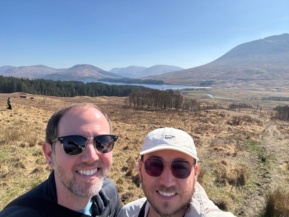 A selfie of two men in front of a vast Highland landscape in Scotland. Behind them, golden-brown grass covers the rolling hills, with a forested patch and a serene lake visible in the distance. Snow-capped peaks rise along the horizon under a clear blue sky. Both men wear sunglasses, and one sports a white cap.