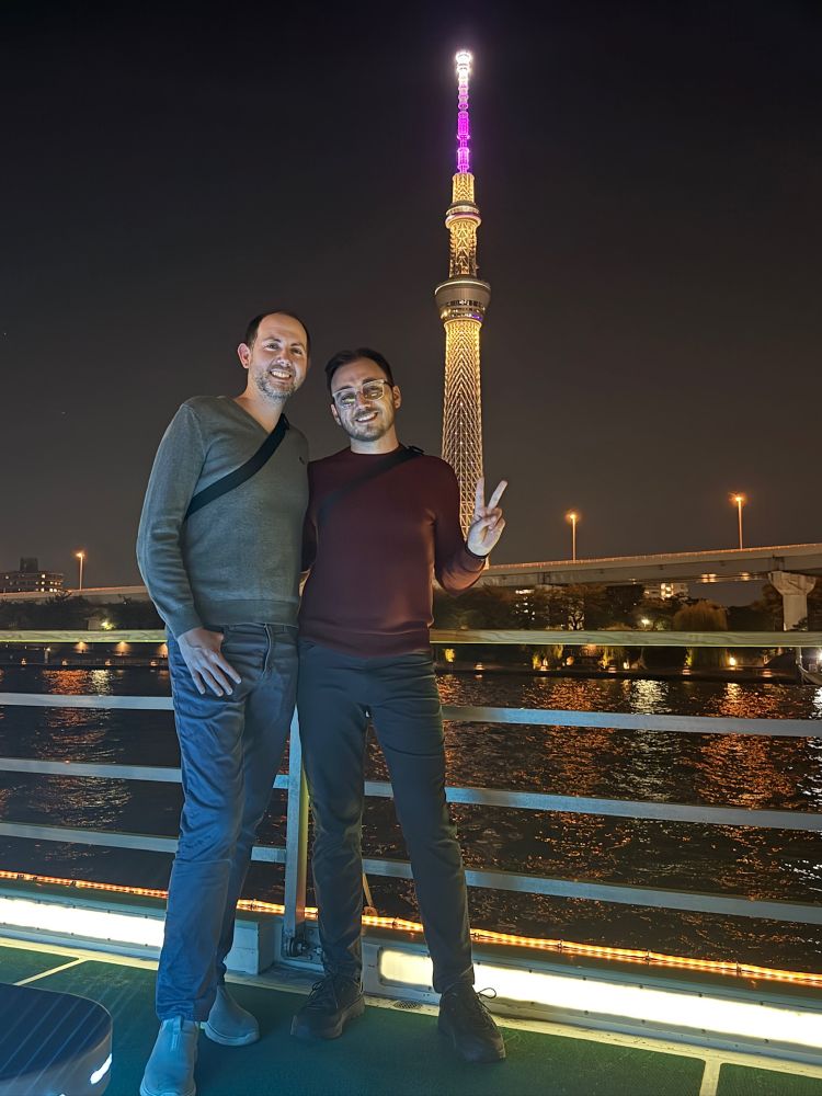 Two men stand smiling on the deck of a boat at night in Tokyo, Japan. Behind them, the Tokyo Skytree rises dramatically into the sky, lit with a gradient of golden yellow and purple lights. The Sumida River sparkles behind them, and a highway bridge stretches across the background. One of the men holds up a peace sign, and both appear warmly dressed for the evening.