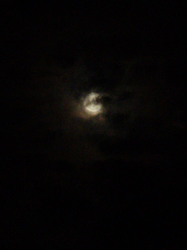 Another photo of the moon, this time only a black background and the blurry glow of the moon shining behind black and charcoal layers of clouds. 