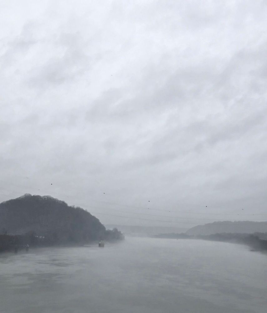 Light fog over the Ohio River, on either side of the river is a small hilly landscape slightly obscured by the fog. 