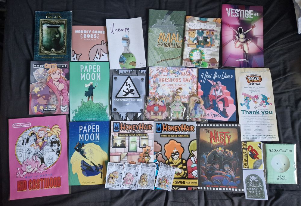 A selection of independent comic books, art prints and keychains lie on a black cloth background. They are arranged in three rows and are of varying sizes, including books of multiple different genres and styles.
