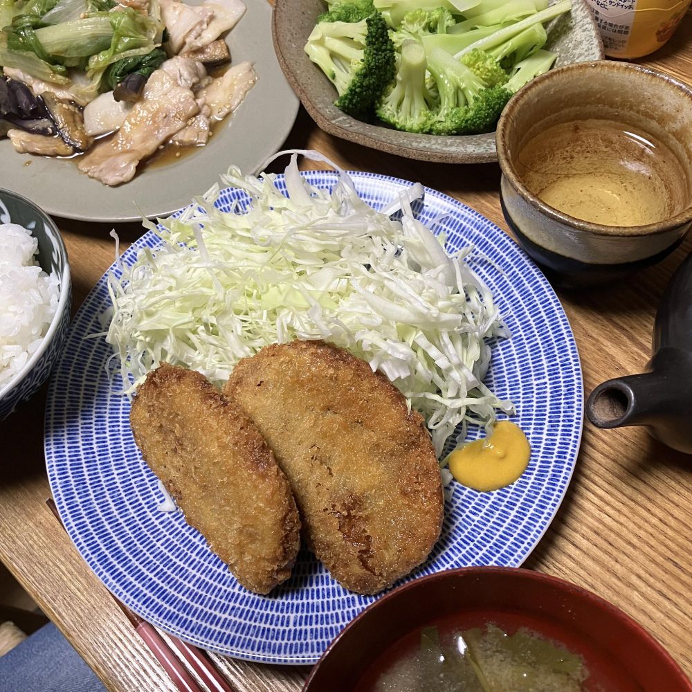 キャベツの千切りとからしを添えたはんぺんフライ2個が青い皿に盛られた和食の食卓。ご飯、味噌汁、炒め物、茹でブロッコリー、湯飲みも並ぶ。