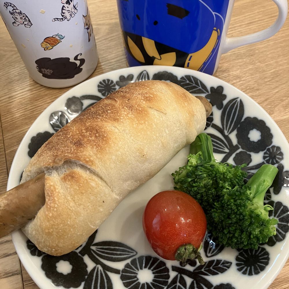 ソーセージ入りのパンとブロッコリー、ミニトマトが白黒の花柄の皿に盛られており、背景には猫柄と青いキャラクターのマグカップが置かれている。