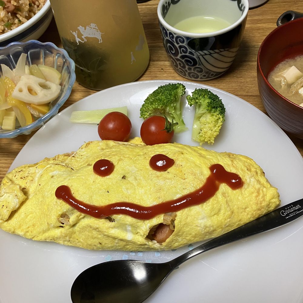 オムライスの上にケチャップで笑顔の顔が描かれている。付け合わせにブロッコリーとミニトマトが添えられた白い皿に盛り付けられている。周囲には味噌汁、飲み物などが並んでいる。スプーンには「cafe Cinnamon」のロゴが刻まれている。