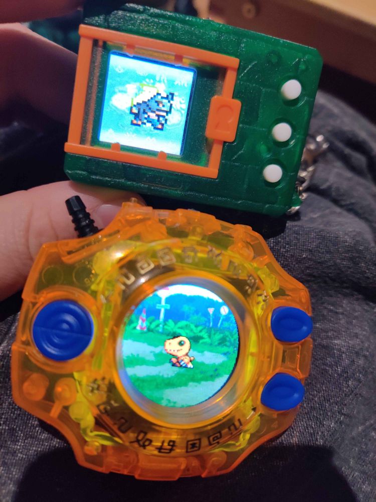A green and orange virtual pet above an orange clear plastic digivice. Both devices are turned on showing pixel sprites of digimon.
