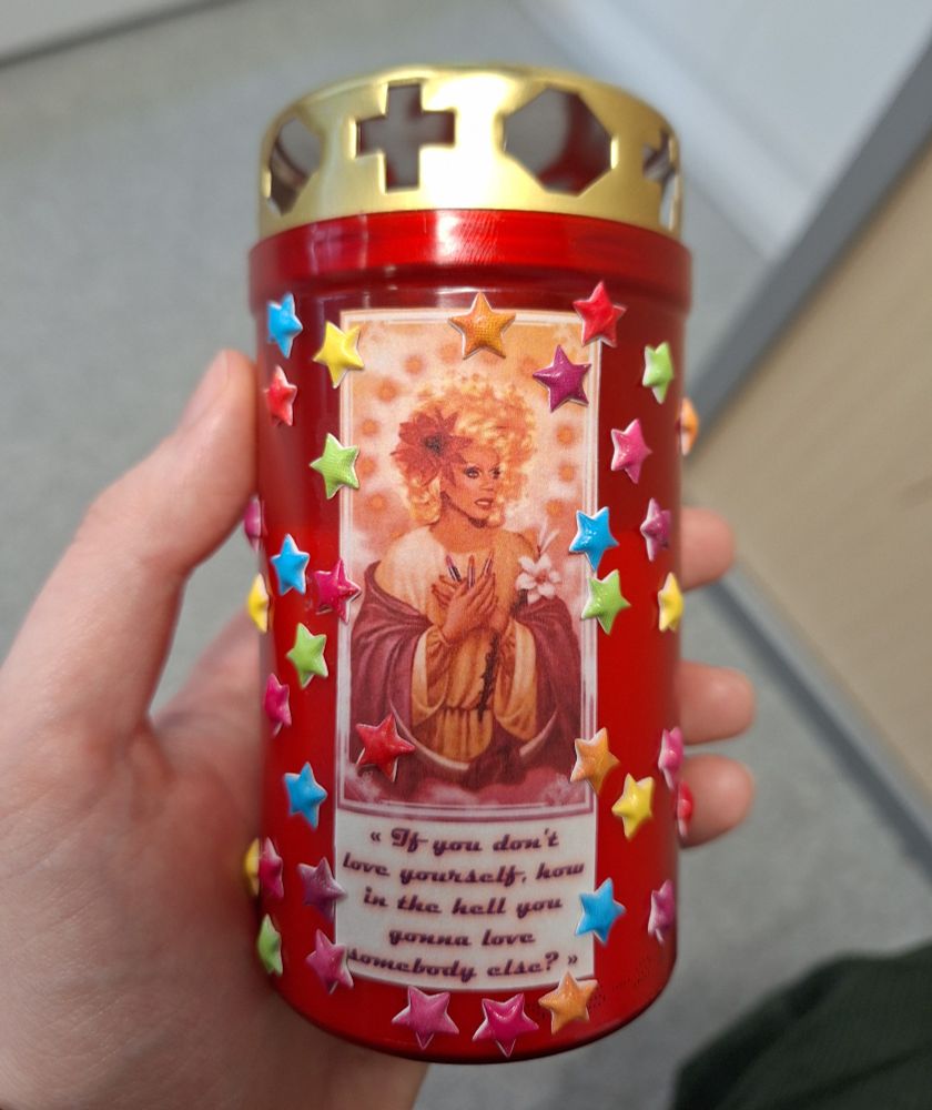 Bougie de cimetière en plastique rouge avec un couvercle doré sur laquelle est collée un montage de RuPaul en Christ, avec la mention "If you don't [sic] love yourself, how in the hell you gonna love somebodey else?". Des petites étoiles en plastique de toutes les couleurs sont collées en farandole sur la bougie autour de l'image.