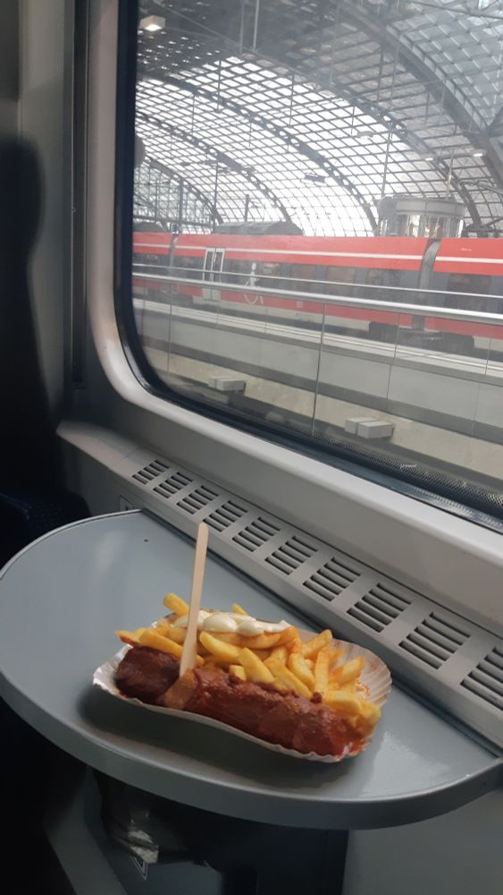 Currywurst vegane et frites dans le compartiment d'un train polonais. À travers la fenêtre, on voit un train InterCity rouge allemand, et la verrière arrondie de l'étage supérieur de la gare centrale de Berlin