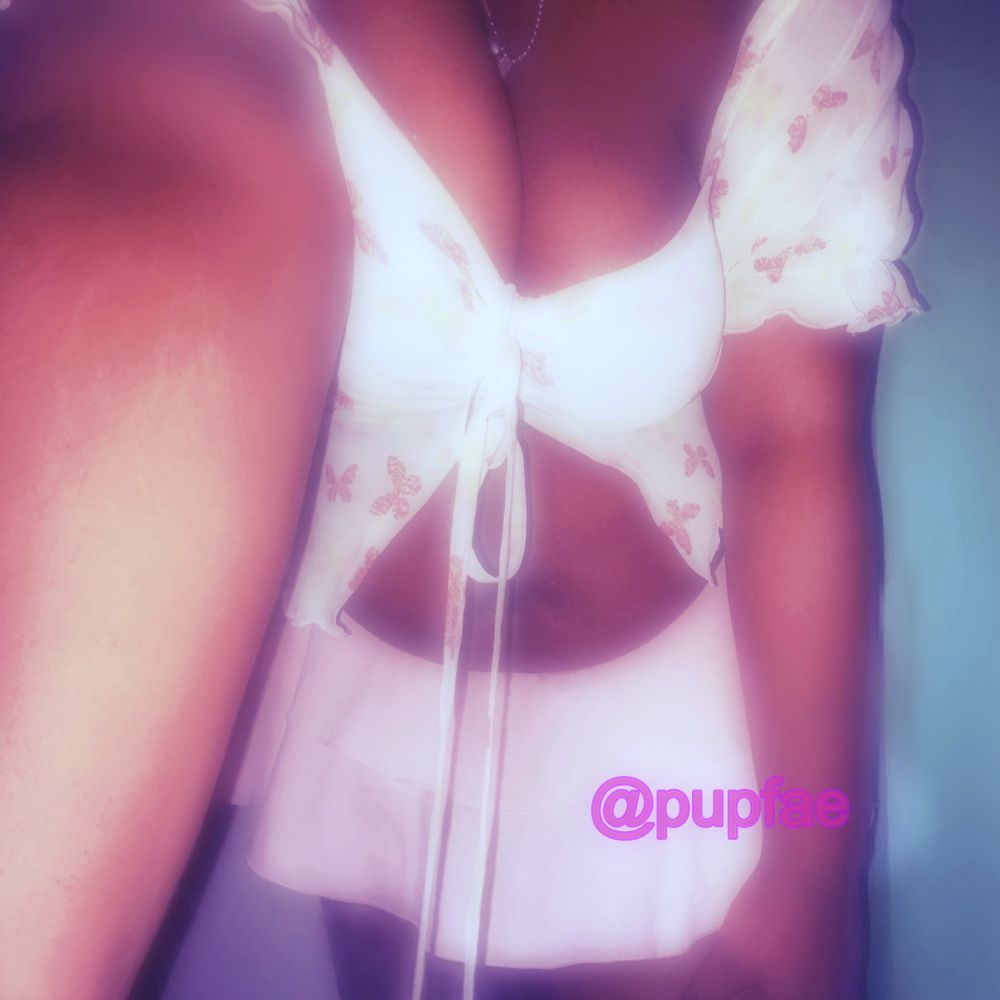 a black femme stands against a blurred blue background, bent over, one hand on his thigh, the other . he's wearing a floral crop top and a pink miniskirt, the crop top showing cleavage. on his thigh is a watermark reading " @pupfae " END ID