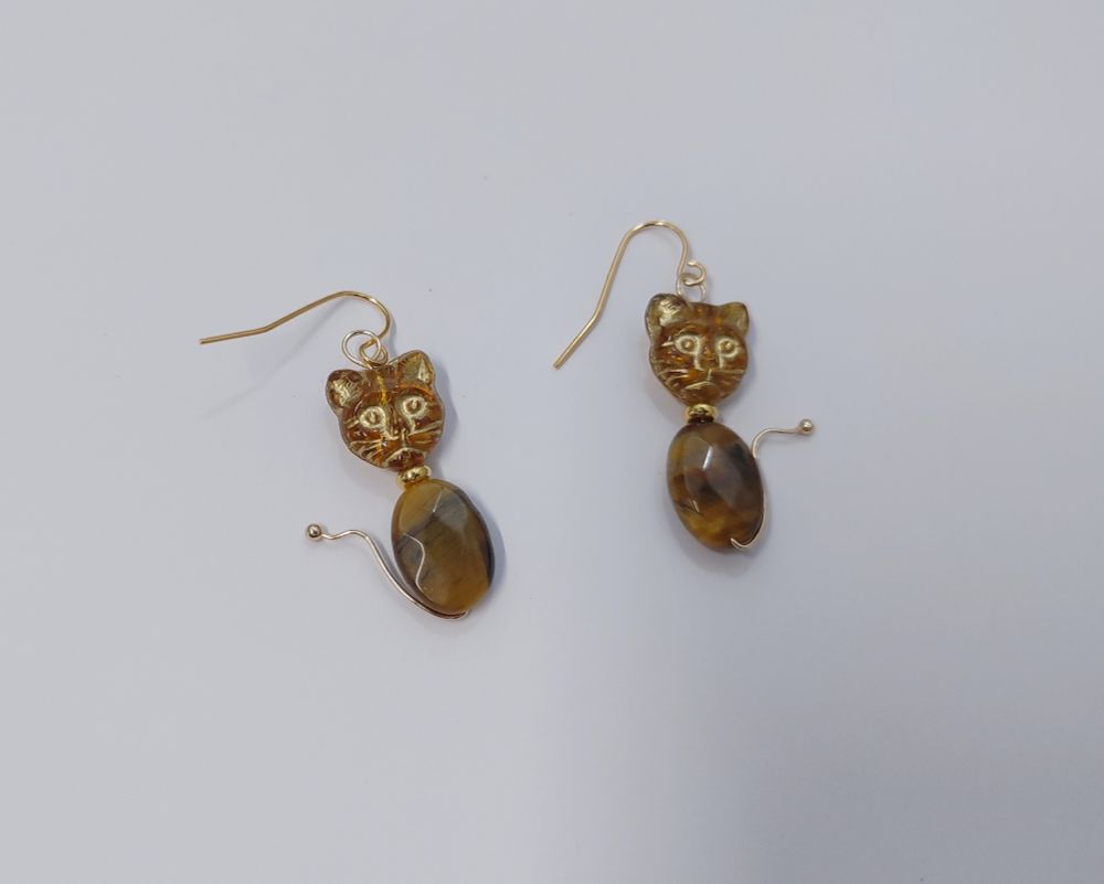 A pair of cat face and tiger's eye earrings lay on a white background. The cat face is made from gilt, transparent, light brown Czech glass. A faceted, 14mm tiger's eye oval functions as the cat's body. The cat is mounted on an 18K gold-plated ball pin that is shaped around it to form a tail. The earring hooks are 22K gold-plated earring wires.