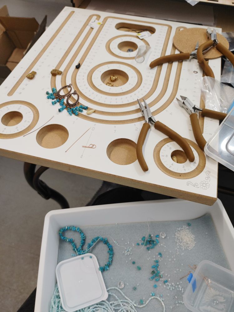 A large bead board on a table and a bead tray on a lap. The board is covered with jewelry-making tools, some cat beads, pieces of copper, a pair of incomplete turquoise chandelier earrings earrings. The bead tray has a mix of loose and stranded turquoise gemstone chips, a hank of hex seed beads, crystal seed beads, and an empty mini tupperware container. 