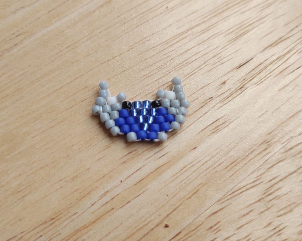 A small, beaded approximation of a blue land crab against a light wood background. The carapace is two shades of blue with a light, shiny blue at the center and a darker blue on the outer edges. It has tiny black eyes and gray claws. 