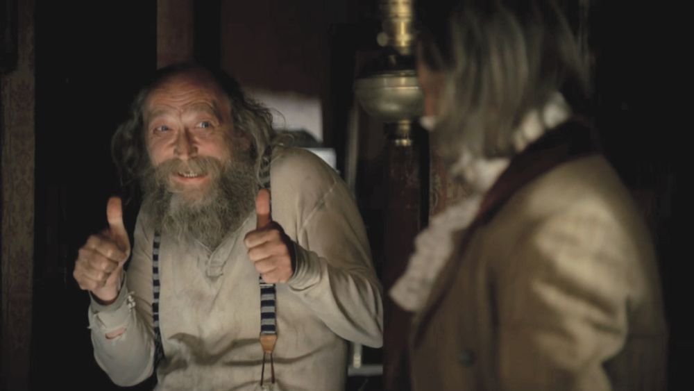 A screenshot from Deadwood showing the character Richardson performing a double thumbs-up gesture while grinning with innocent delight.