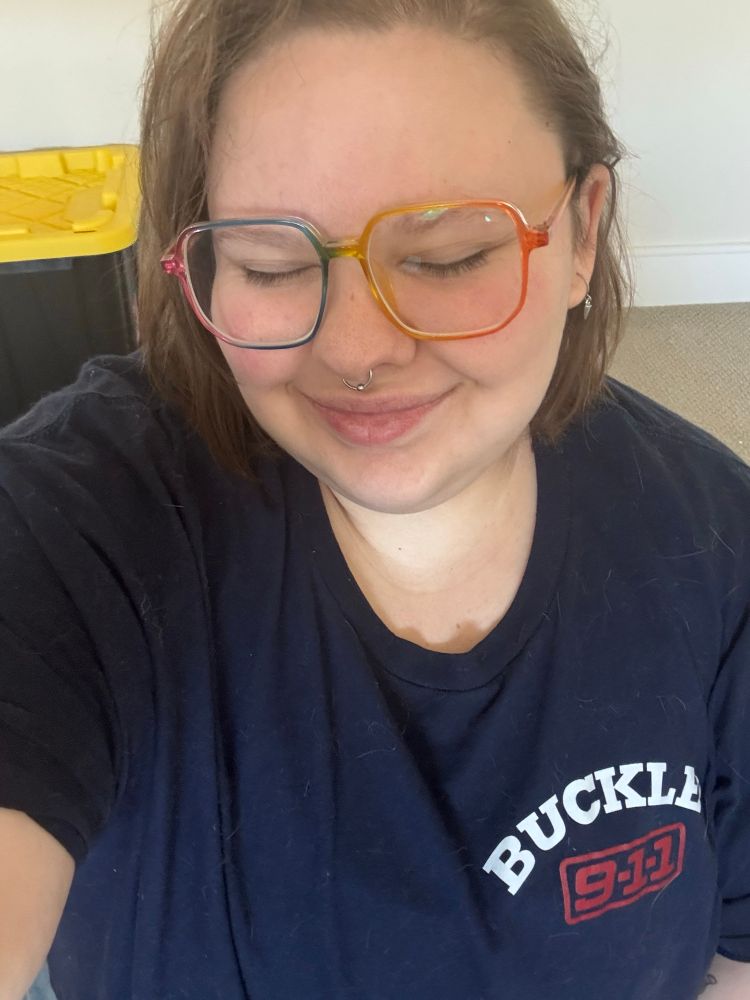 campbell taking a selfie, wearing a blue buckley 9-1-1 shirt and grinning with their eyes closed 