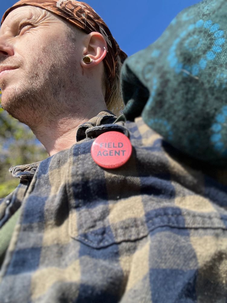 Upward photo of my green checkered shirt sporting a 1" red Field Agent button badge with my face in the background looking off into the sunny distance