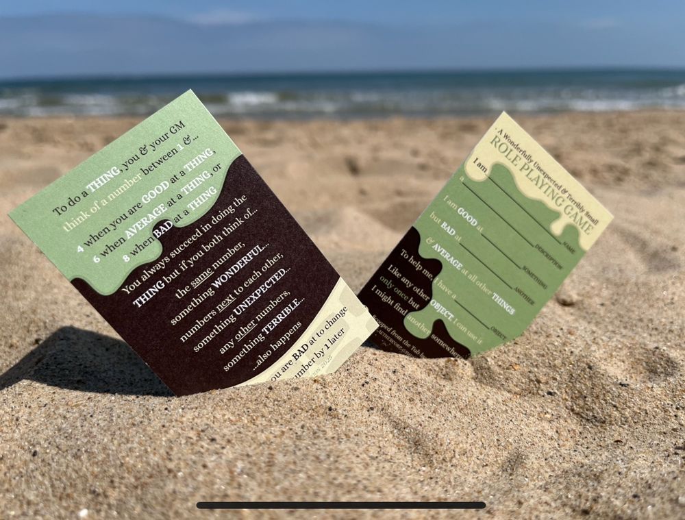 Front and back of A Wonderfully Unexpected & Terribly Small RPG stuck in sand at a beach 