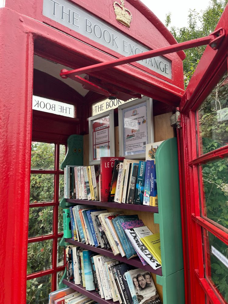 A community book exchange in a red telephone box with a copy of Orchidelirium, a copy of Advanced Double D6 and two Field Agent Handbooks nestled on one of the shelves