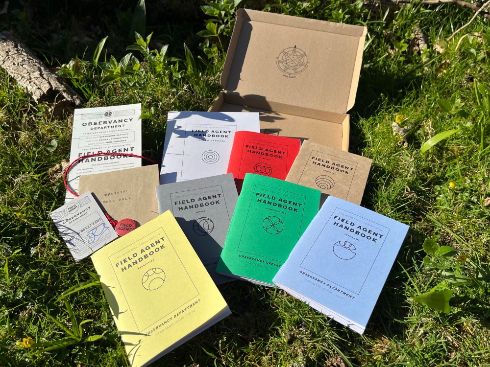 A whole set of Field Agent Handbooks with ID card, button badge, handbill, red string with key and a red button badge spread out on a grassy mound