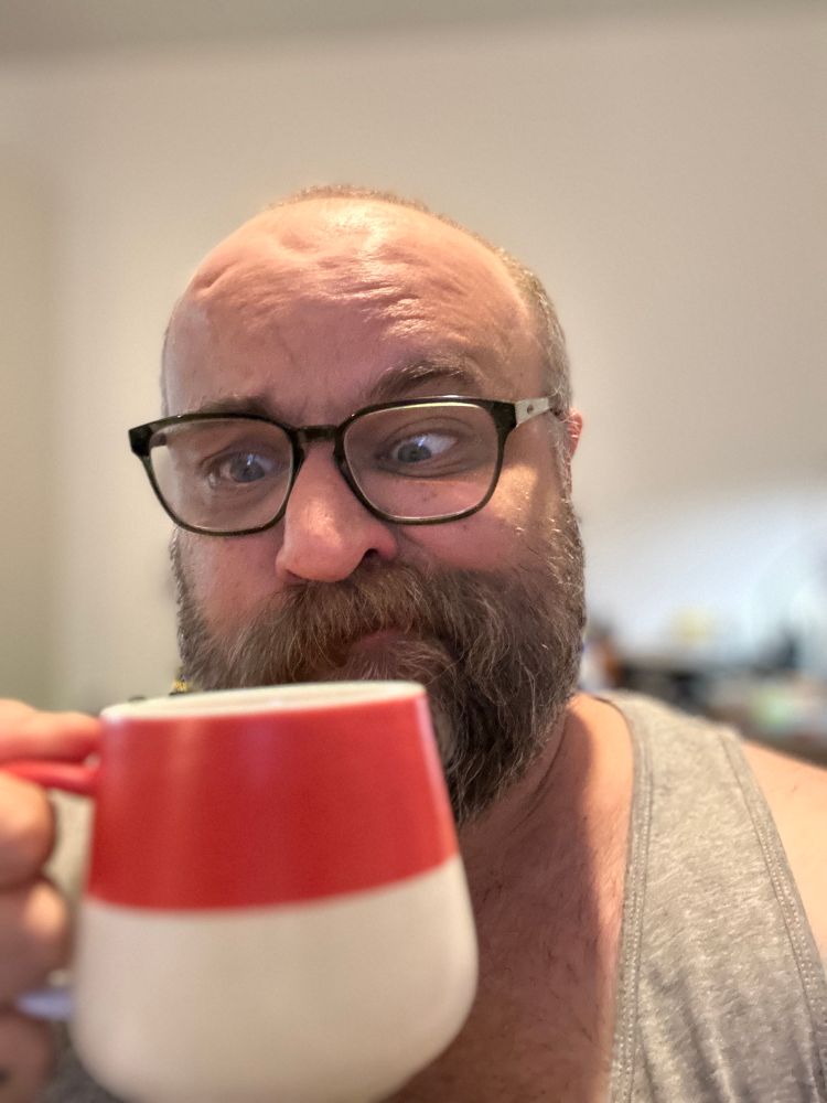 Photo of a bald, bearded, bespectacled Roma man in a grey vest staring in love and sad desperation at a mug of tea