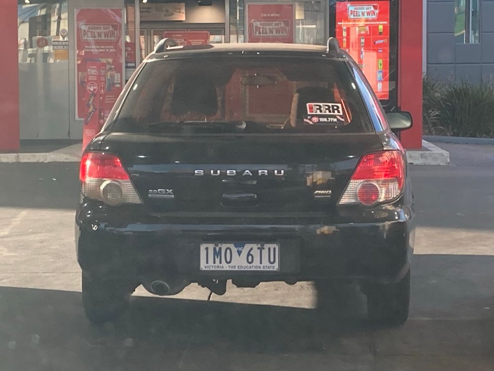 A black Subaru at a petrol station with both a 3RRR and a 3PBS sticker on the lower right hand side of the rear window. 