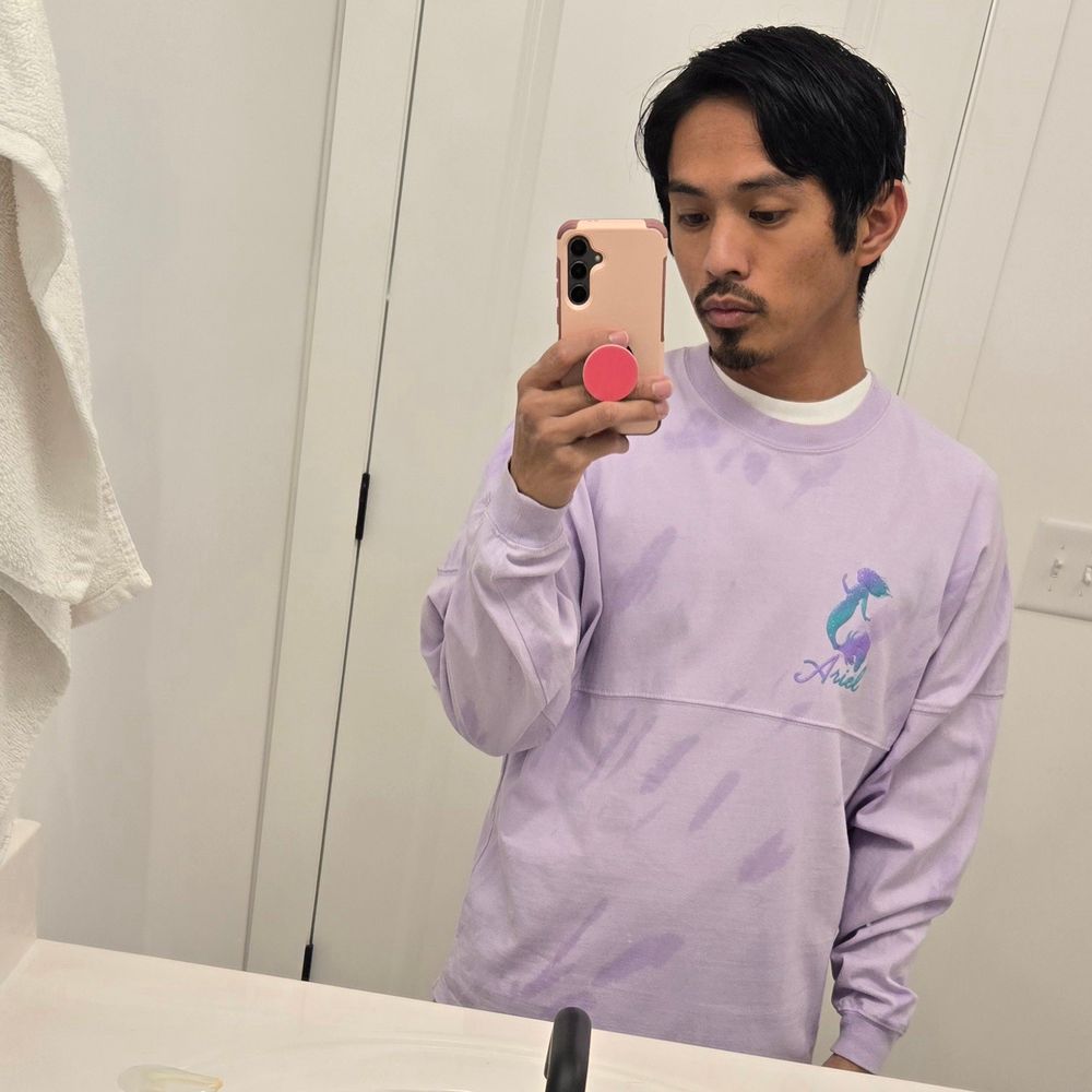 Selfie I took in an all white bathroom. I am wearing a light purple sweater with a silhouette of Ariel from Disney's live action "The Little Mermaid" on the chest. Beneath the image is cursive text that reads "Ariel". The silhouette and text is colored to be a deep purple and teal gradient.