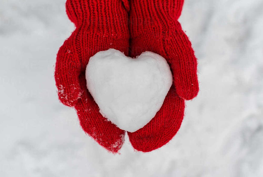 A pair of hands in wooly red mittens shown holding a nicely packed snowball in the shape of a heart above the snow-covered ground.