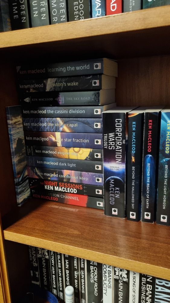 Photo of part of a book shelf showing the spines of books by Ken Macleod stacked horizontally and vertically.