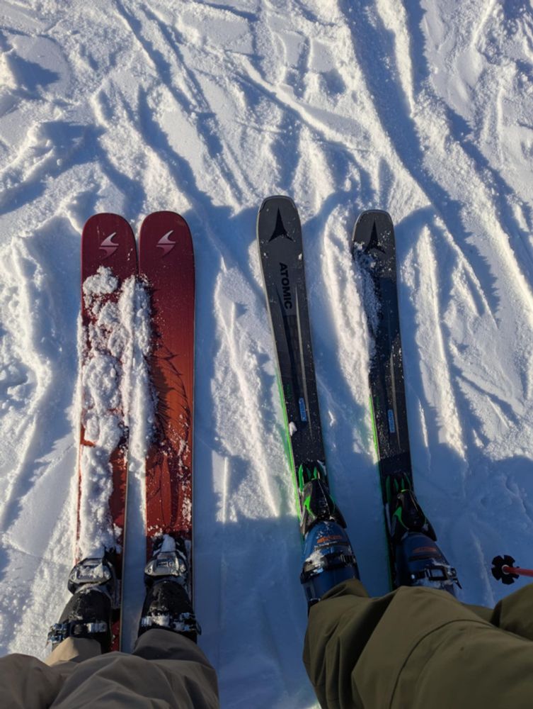 Photo of the tips of two pairs of skis with skiers legs and boots taken from above on snow.