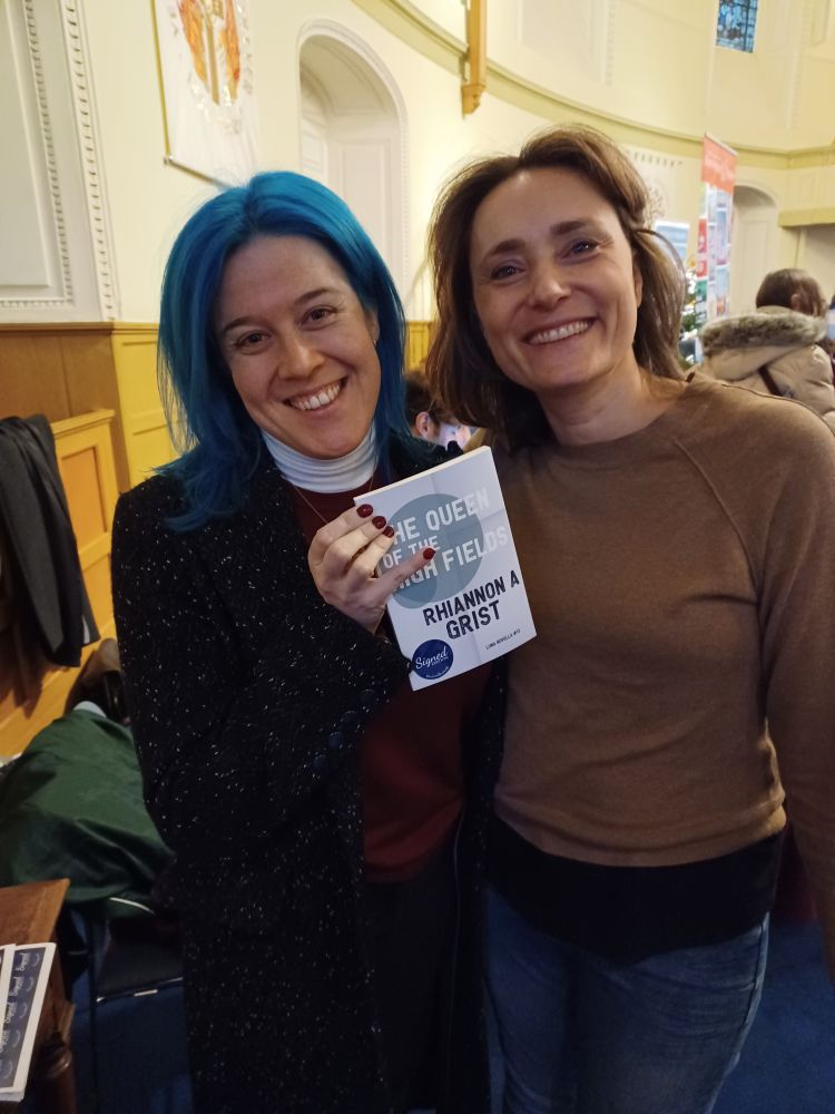 Rhiannon and Fra pose together as Rhiannon holds up a copy of her award-winning novella with a "signed" sticker on it.