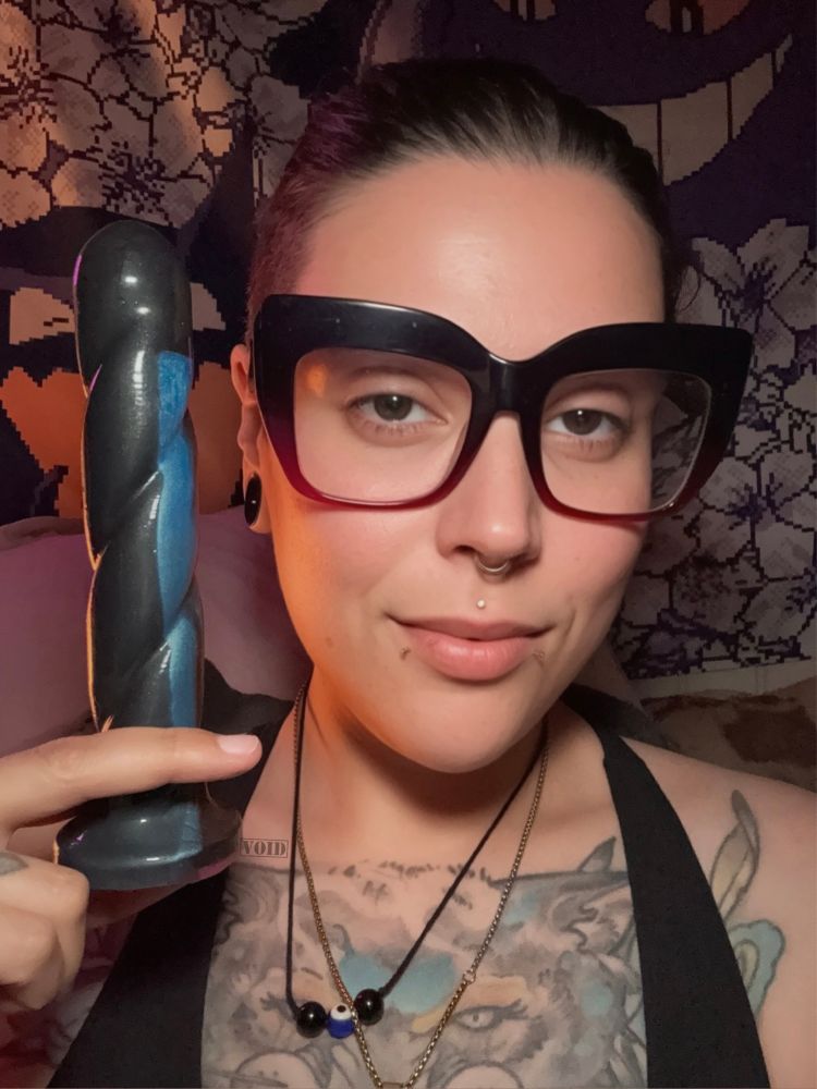Void holding a no frills dildo by Funkit Toys that is twisty and multi colored. Void is wearing a sports bra and is smiling holding the dildo up. 