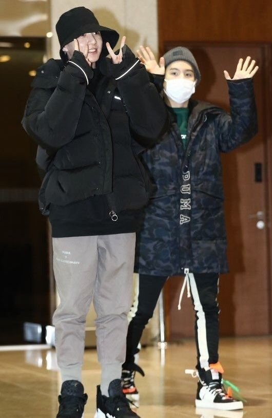 Yoonkook at the airport waving and giving peace signs for the camera. They are dressed in winter jackets and hats with sweats underneath.