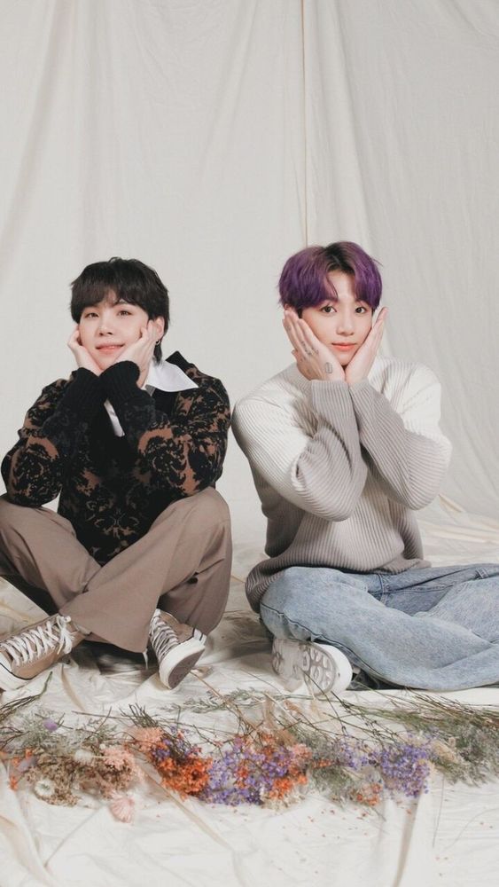 Yoonkook sitting in front of a flowy white background posing like aegyo flowers. There are flowers laid at their feet and they are wearing sweaters and Chucks.