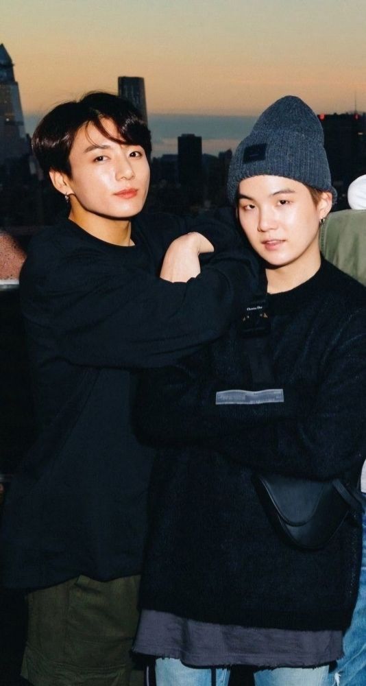 Yoonkook in dark colors on a rooftop at sunset. They both have their arms crosses, JK's resting on Suga's shoulder, and Suga has on a pointy gray beanie.