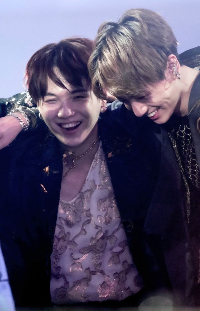 Blonde Jungkook with his arm around Suga's shoulders as they perform on stage, laughing and smiling.