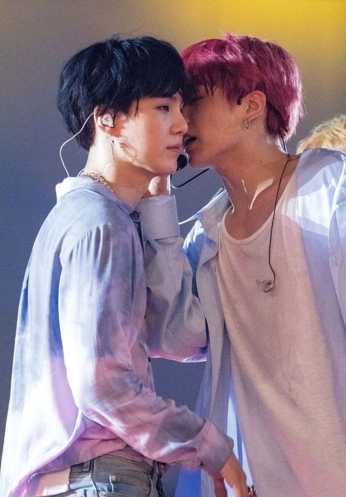 Cherry haired Jungkook whispering into Suga's ear while they are on stage in light pastel clothing.