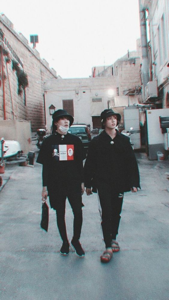 Yoonkook in all black casual clothing and buckets hats wandering in an alley, looking a bit unsure. 