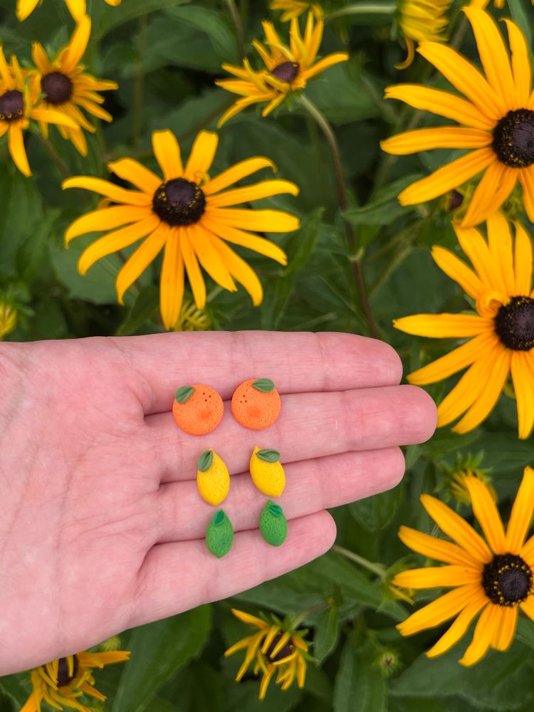 Three pairs of stud earrings on my palm against a background of blooming Black Eyed Susan flowers. The top design is an orange with a little bit of shading and a leaf; the middle design is a lemon with a leaf; the bottom design is a lime with a bit of shading and a leaf at the top.
