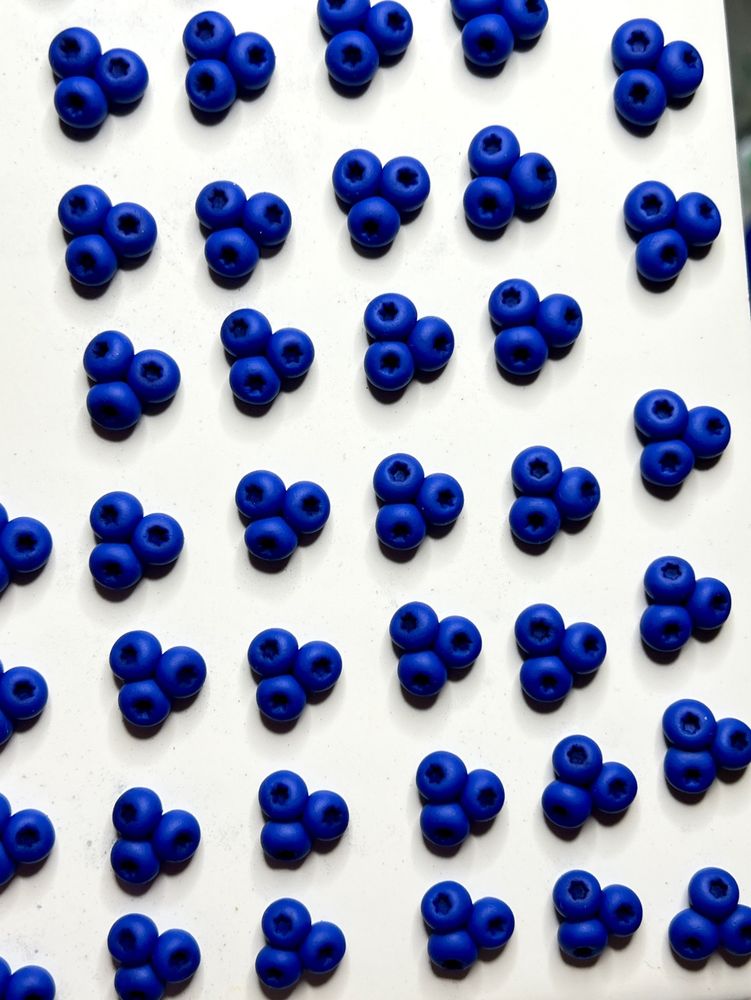 A white ceramic tile full of small clusters of polymer clay blueberries. Each cluster has three berries that are a deep blue, with a divot with a darker purpley-blue color as shading.
