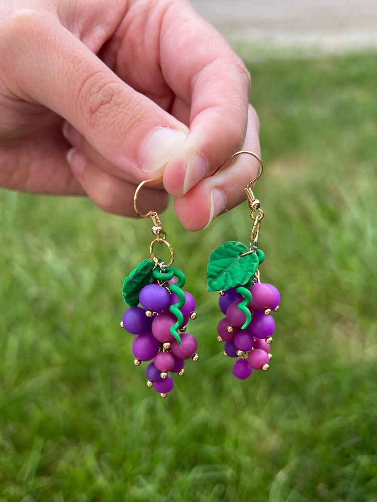 A pair of purple grape earrings held up against a background of grass. There are three shades of reddish purple grapes, a green vine, and a green grape leaf. There are metallic gold balls at the bottom of each grape that hold the piece together.