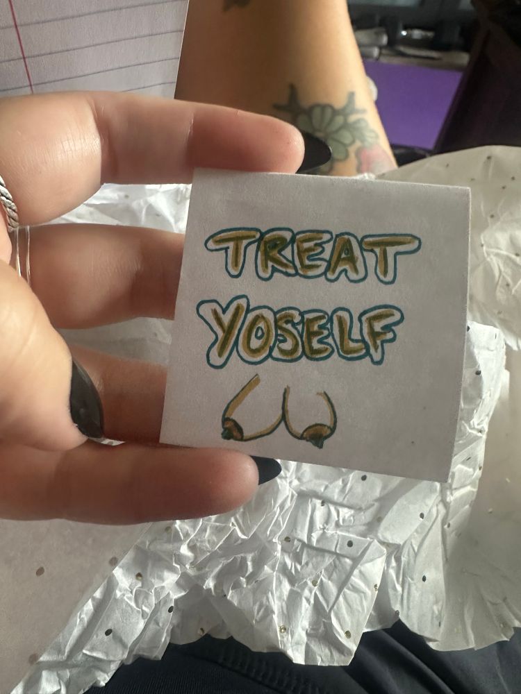 Amy’s hand holding a note that says treat yourself with a doodle of tits underneath it 