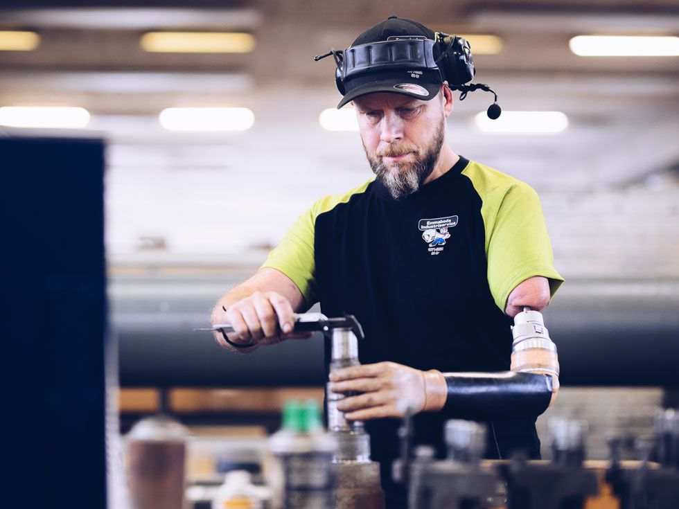 A man with a prosthetic arm connected directly to his upper arm via a titanium implant extending through the skin. He is working in a machine shop, holding a part with his prosthetic hand and measuring its diameter with a set of calipers in the other hand