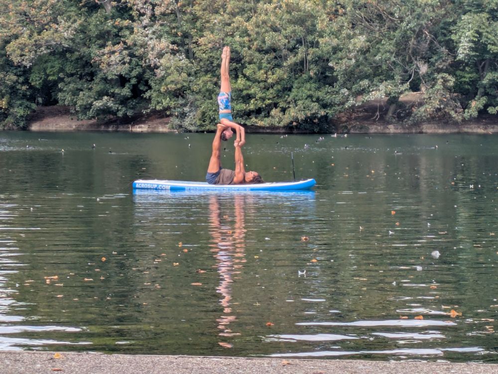 A woman and man perform gymnastics on an inflatable paddle board in the middle of a lake.  The man is lying on his back with his legs at 90 degrees. The woman is in a handstand position, with her shoulders on the man's feet and holding his hands to help balance. 